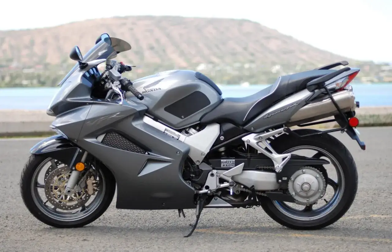 Szary motocykl Honda VFR 800 z widokiem na wodę i wzgórze w tle.