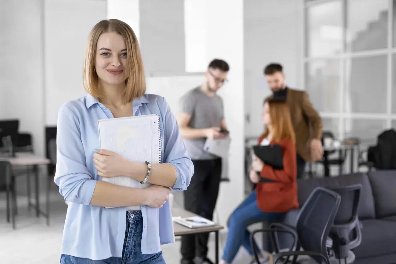 Uśmiechnięta kobieta z notatnikiem, w tle zespół dyskutujący o możliwościach, jakie daje dofinansowanie z urzędu pracy.