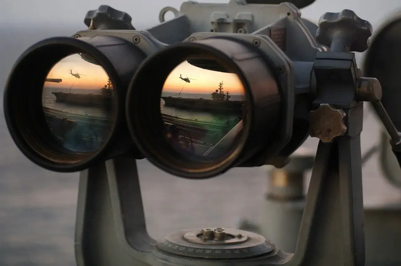 Fernglas-Werte: Ein Flugzeugträger und ein Hubschrauber spiegeln sich im Okular eines Fernglases bei Sonnenuntergang.