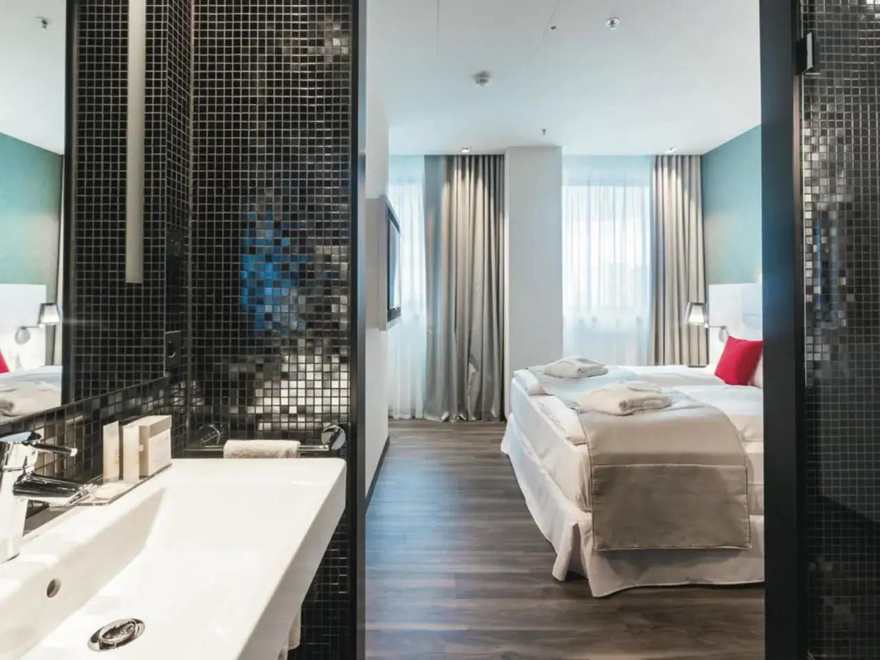 Badezimmer mit Waschbecken und Blick auf ein Hotelzimmer mit Bett und Fenstern.
