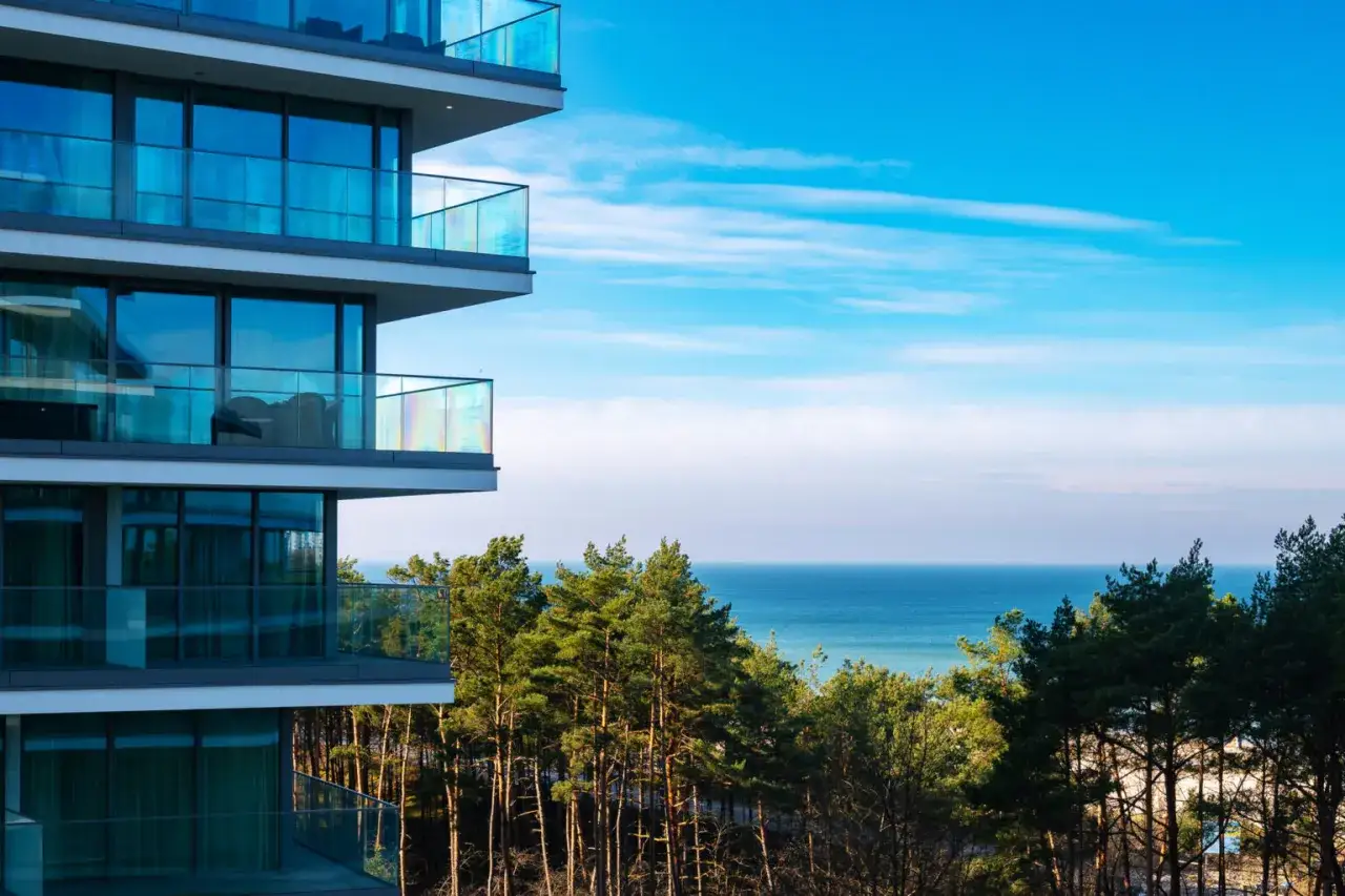 Nowoczesny budynek z balkonami i widokiem na morze. W oddali las i błękitne niebo. Idealne miejsce na wypoczynek w Wave Międzyzdroje Resort&SPA.