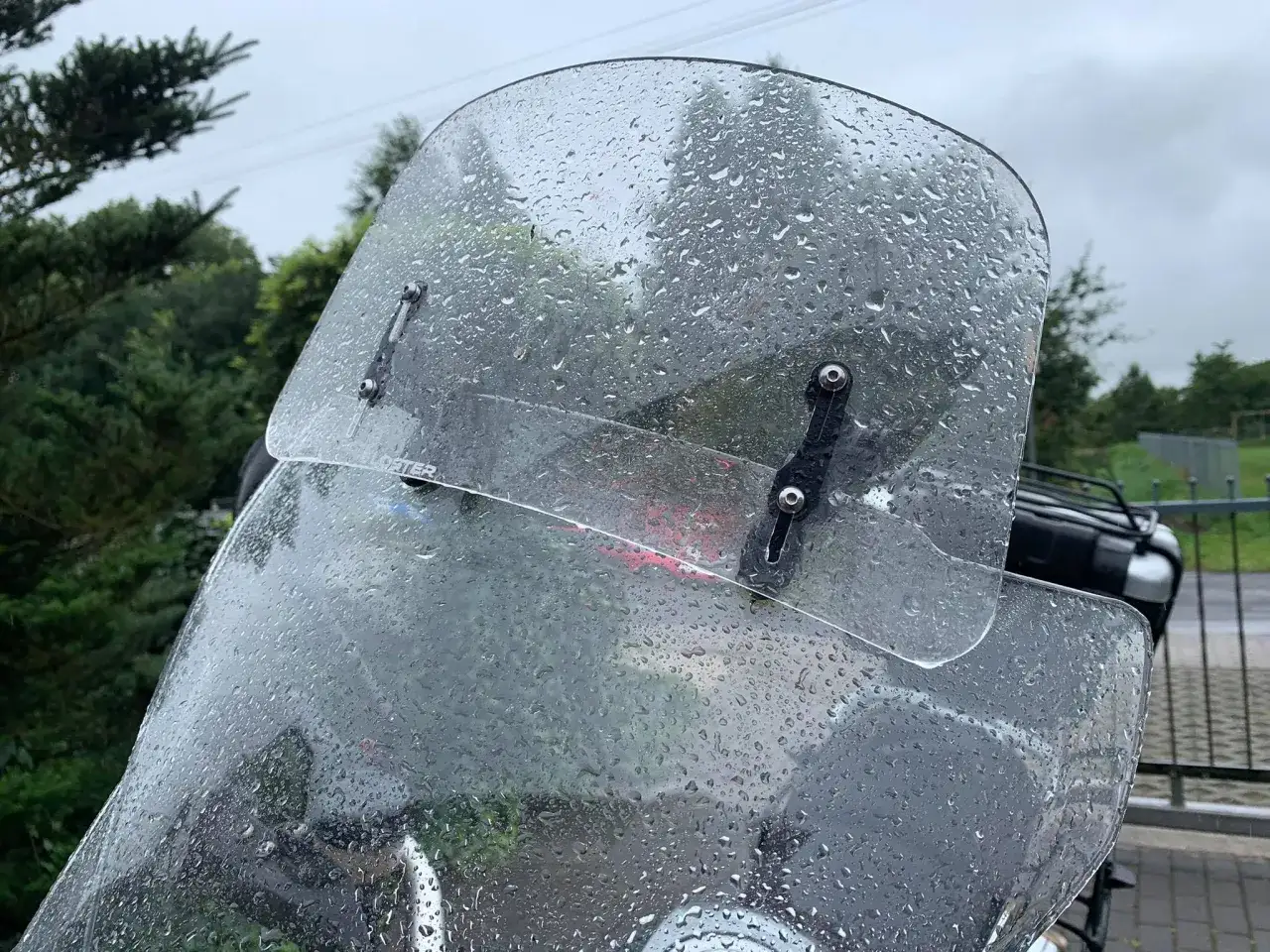 Przednia szyba motocykla pokryta kroplami deszczu, z widocznymi mocowaniami.