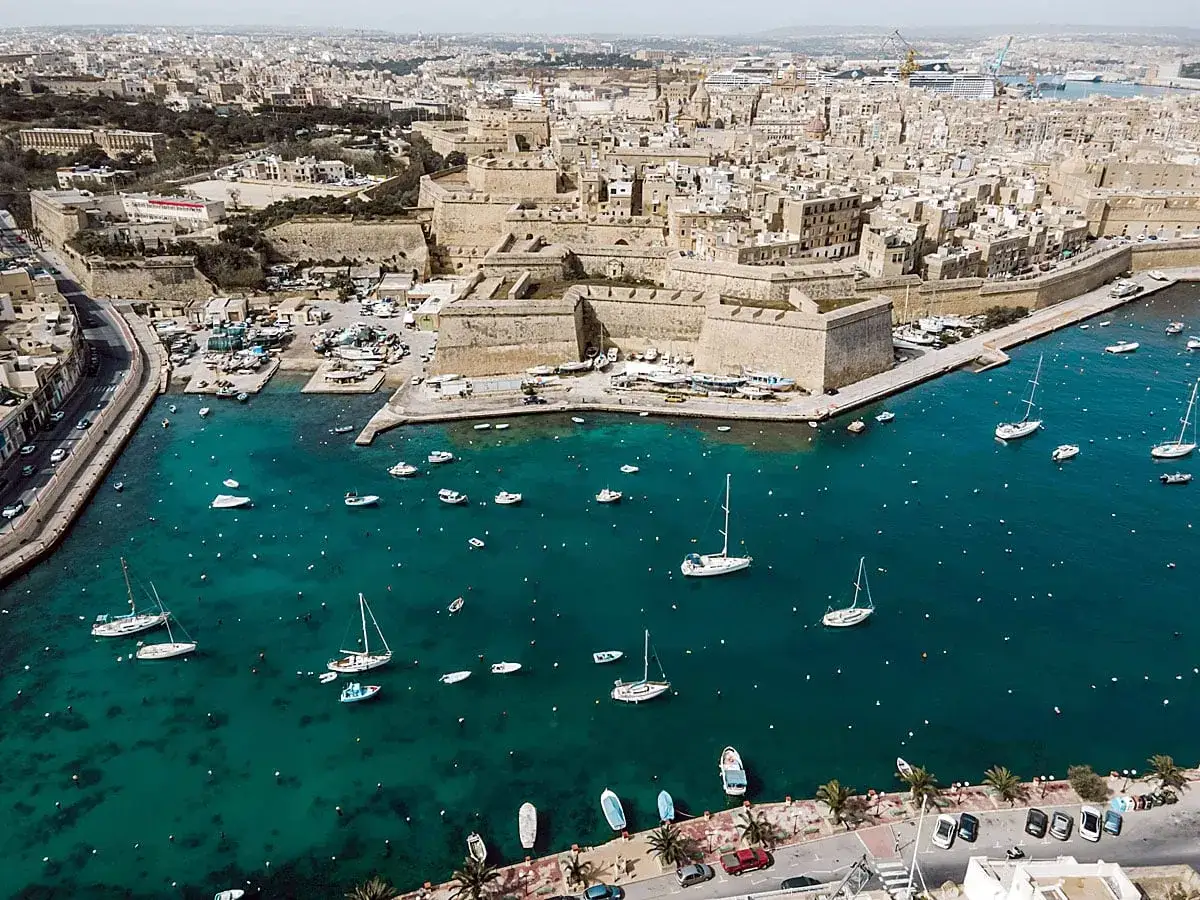 Segeln in Malta: Boote und Yachten im türkisfarbenen Wasser vor einer historischen Stadtkulisse mit Festungsmauern.