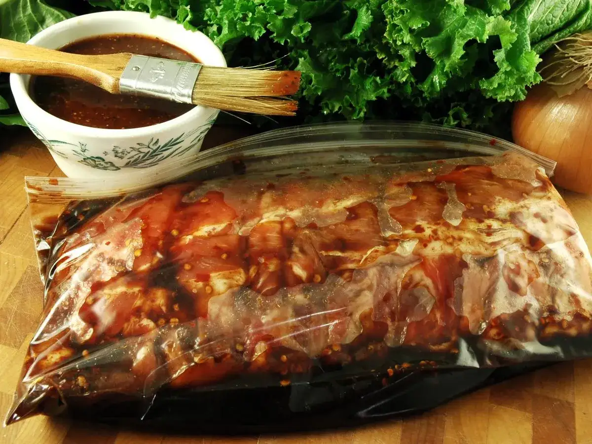 Karkówka w torebce, gotowa na grilla. W miseczce czeka aromatyczna marynata do karkówki na grilla, a obok pęczek zielonej sałaty i cebula.