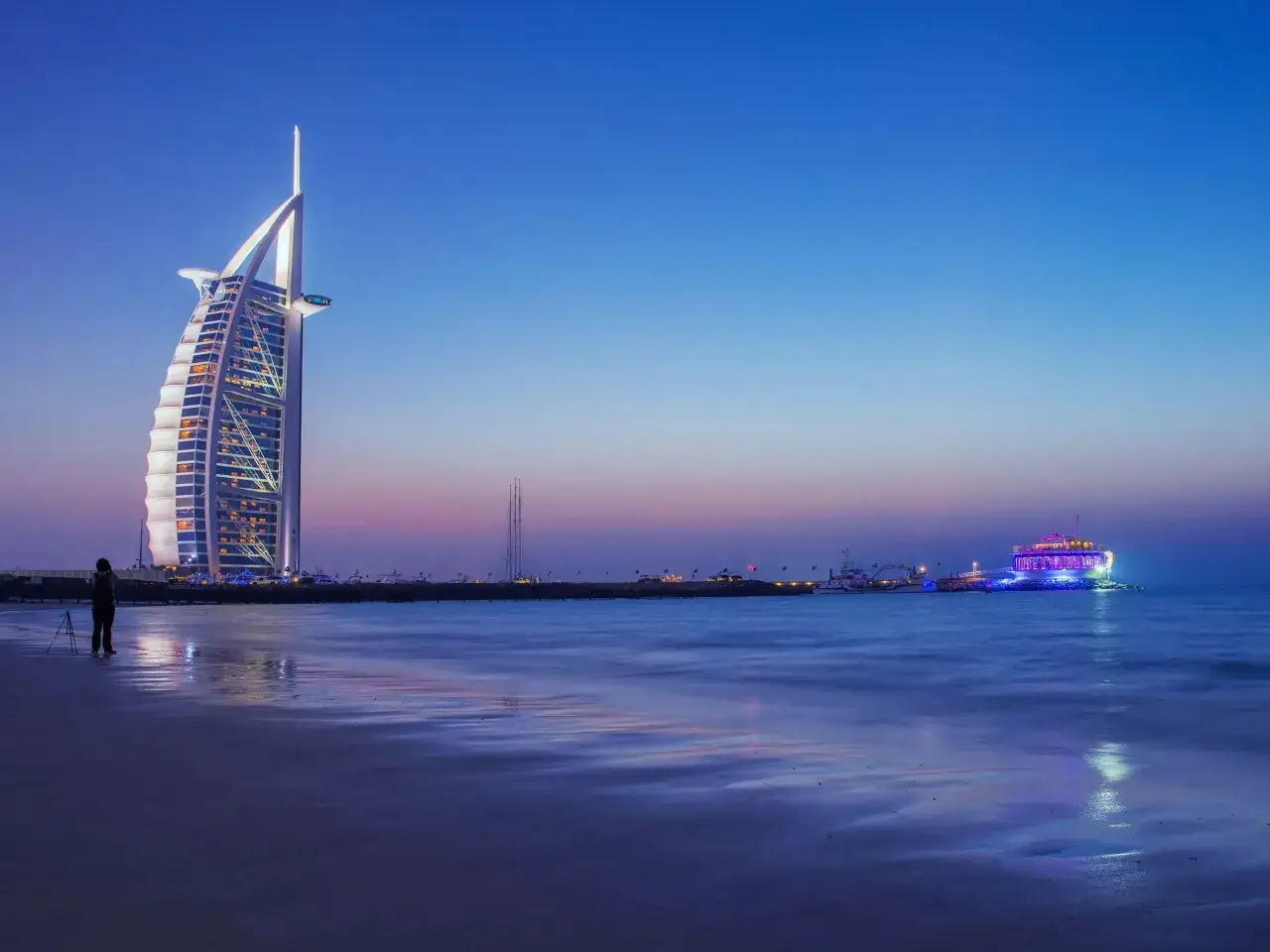 Zachód słońca w Dubaju, z luksusowym hotelem Burj Al Arab i oświetloną łodzią. Zastanawiasz się, ile kosztuje wycieczka do Dubaju?