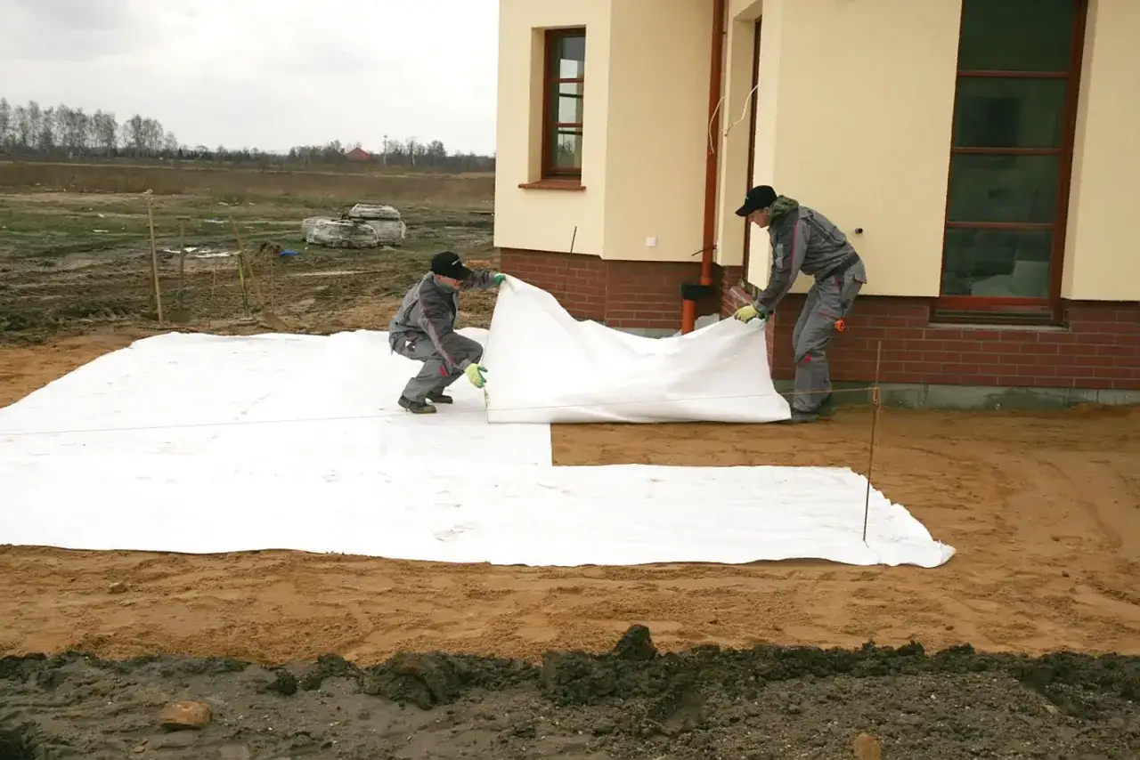 Dwóch robotników rozkłada białą geowłókninę, przygotowując tani taras na gruncie.