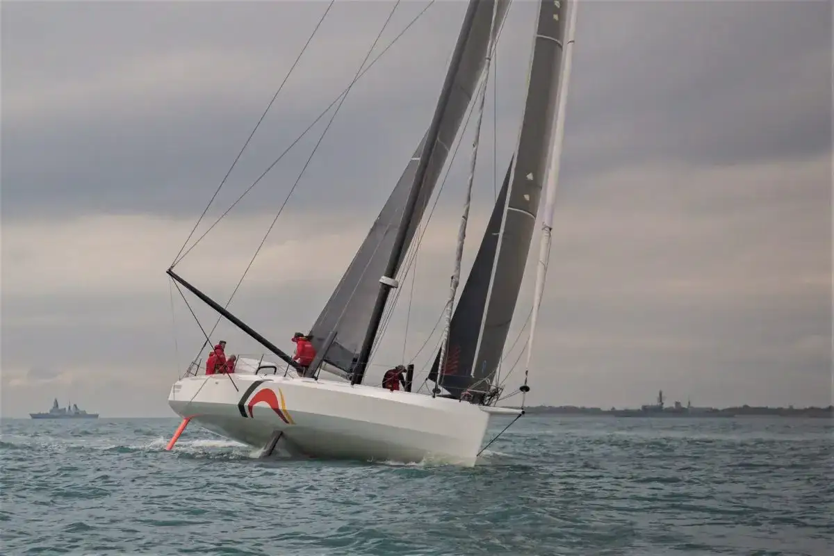 Ein IMOCA 60 segelt auf stürmischer See, die Crew in roten Jacken bereitet sich auf die nächste Welle vor.