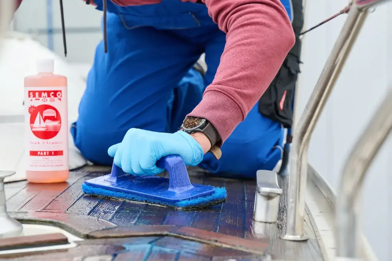 Person reinigt mit blauer Bürste und SEMCO Teak Cleaner das Holzdeck eines Bootes.