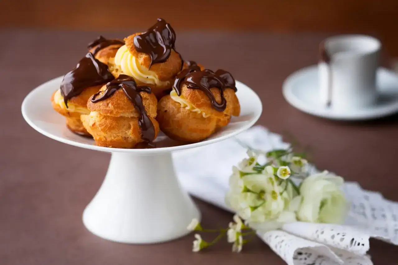 Un plateau blanc présente des profiteroles gastronomiques garnies de crème et nappées de chocolat noir. Des fleurs délicates ajoutent une touche de fraîcheur.