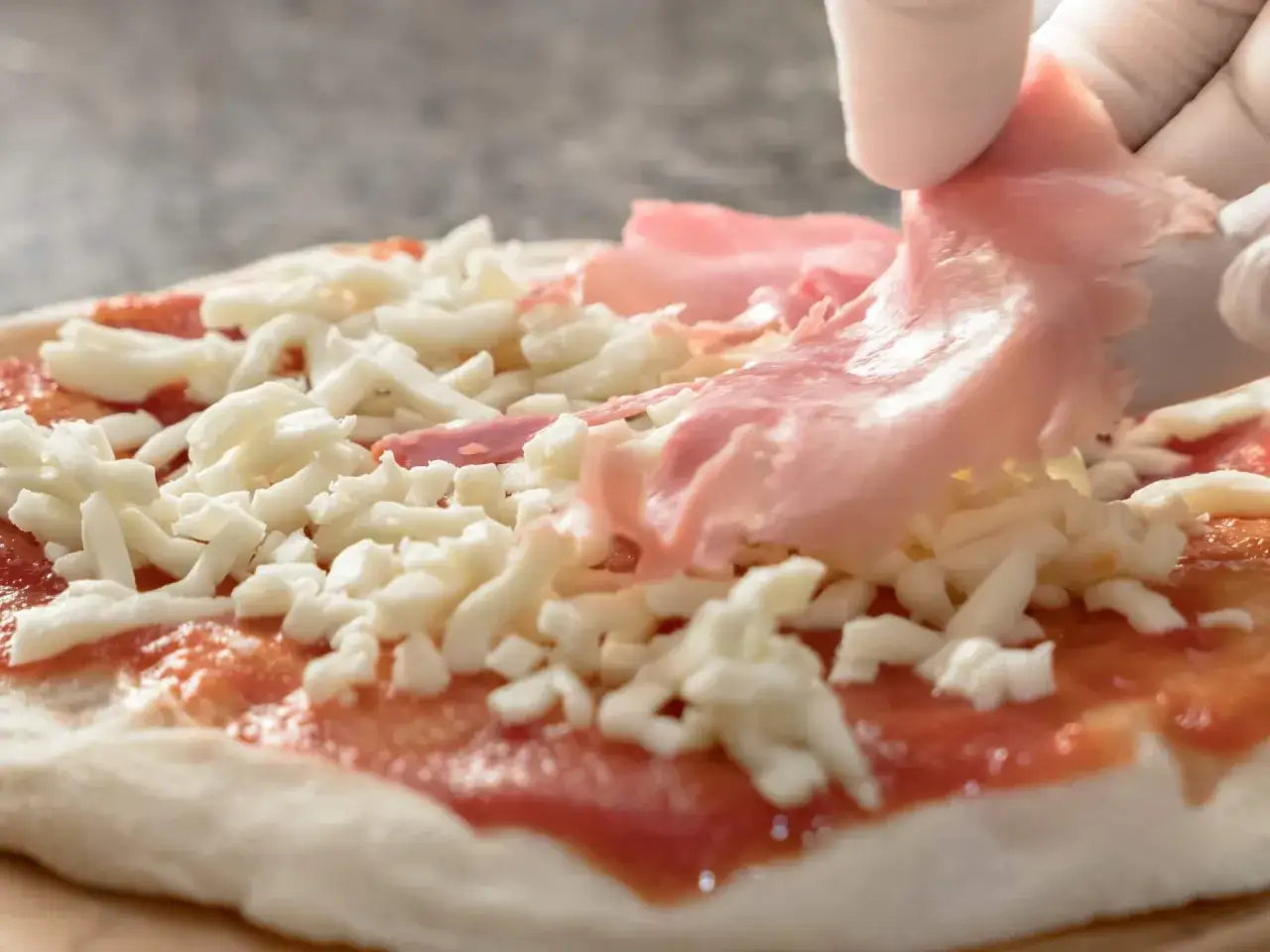 Ręka w białej rękawiczce kładzie plasterki szynki na pizzy z serem i sosem pomidorowym.