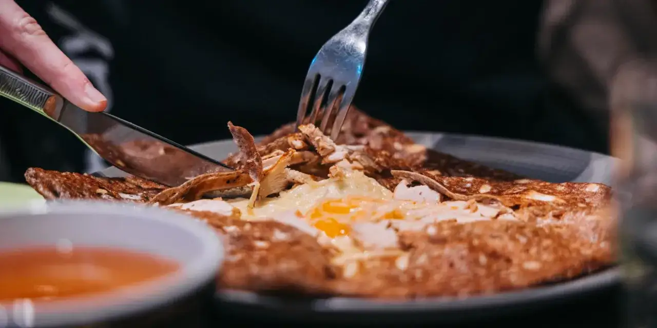 Une galette garnie d'œuf et de poulet, un délice des spécialités de Bretagne, est coupée avec un couteau et une fourchette.