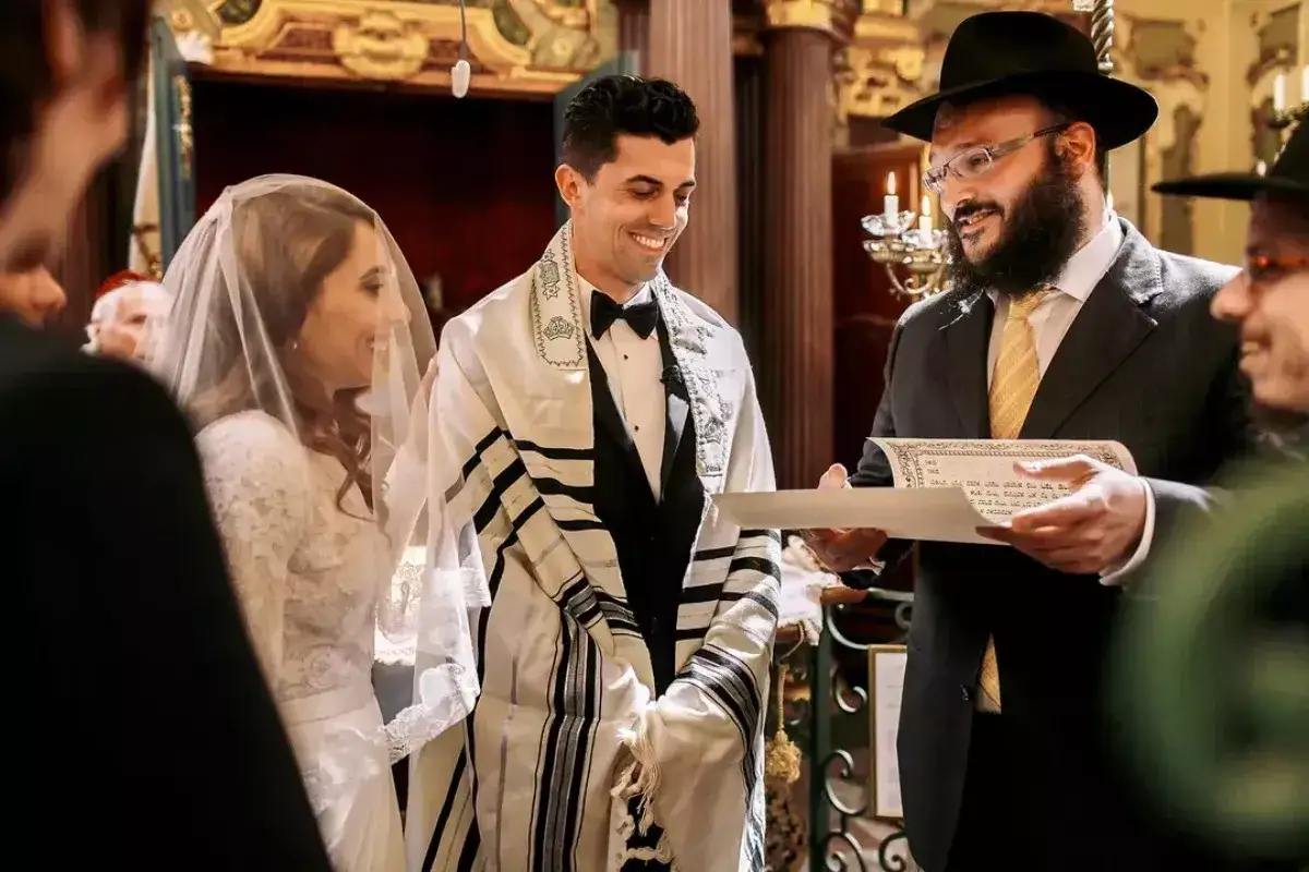 Novios sonríen durante su **matrimonio judío**. El novio lleva un talit y el rabino lee un documento.