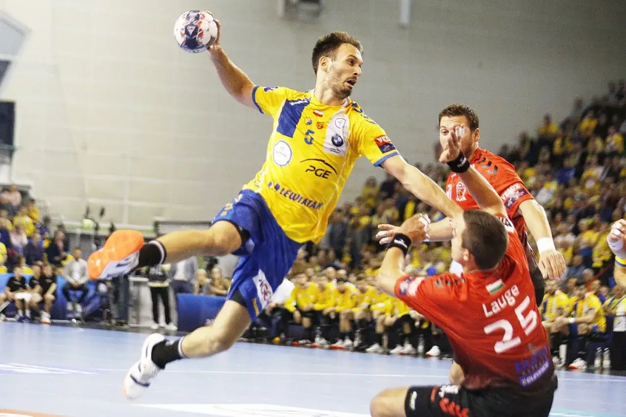 Piłkarz ręczny Telekom Veszprem w żółtej koszulce skacze do rzutu, broniony przez zawodnika w czerwonej.