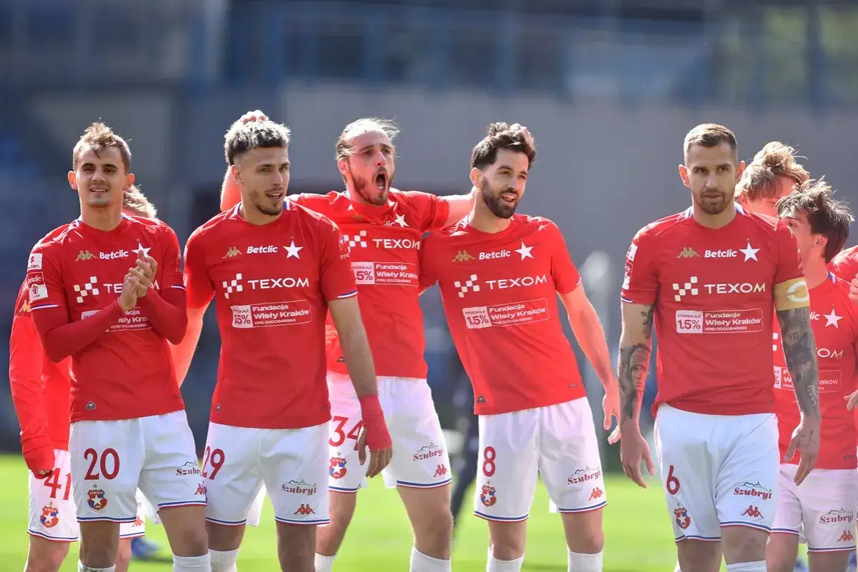 Piłkarze Wisły Kraków w czerwonych strojach, z numerami na koszulkach i spodenkach, świętują. W tle trybuny. #wisla #krakow #transfery