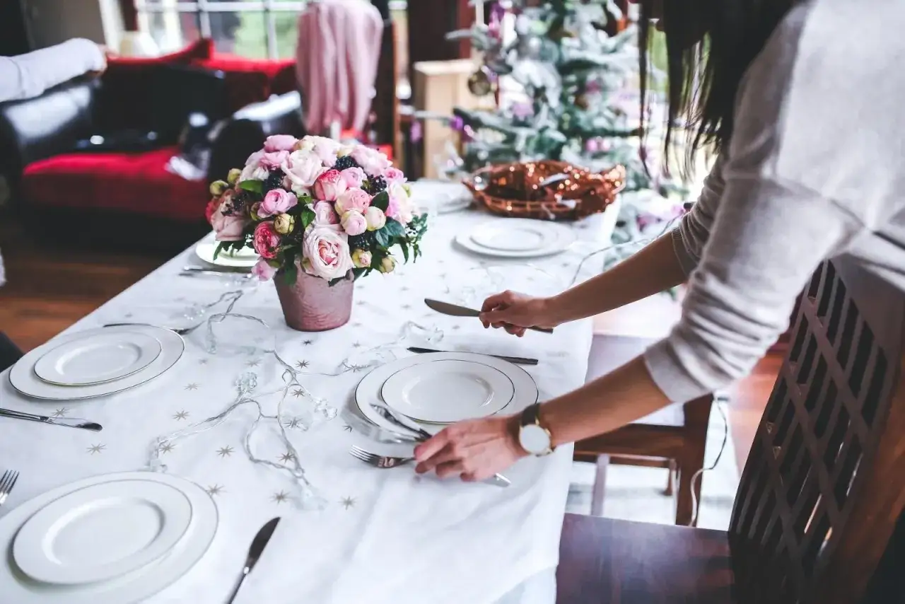 Une table dressée avec un bouquet de roses roses, des assiettes blanches et des guirlandes lumineuses. Une femme ajuste les couverts, ajoutant un caractère élégant à la scène.