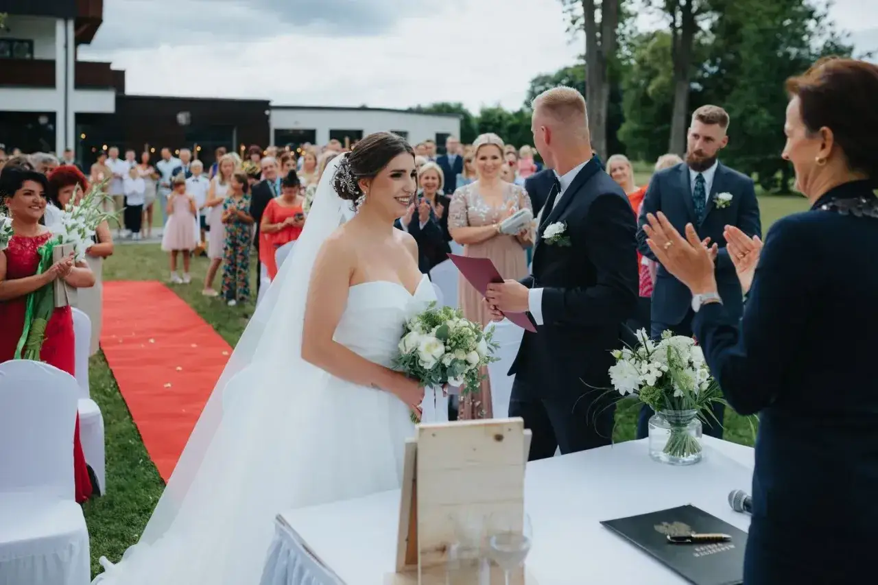 Para młoda podczas ceremonii ślubu cywilnego na zewnątrz. Panna młoda w sukni, pan młody w garniturze. Goście obserwują.