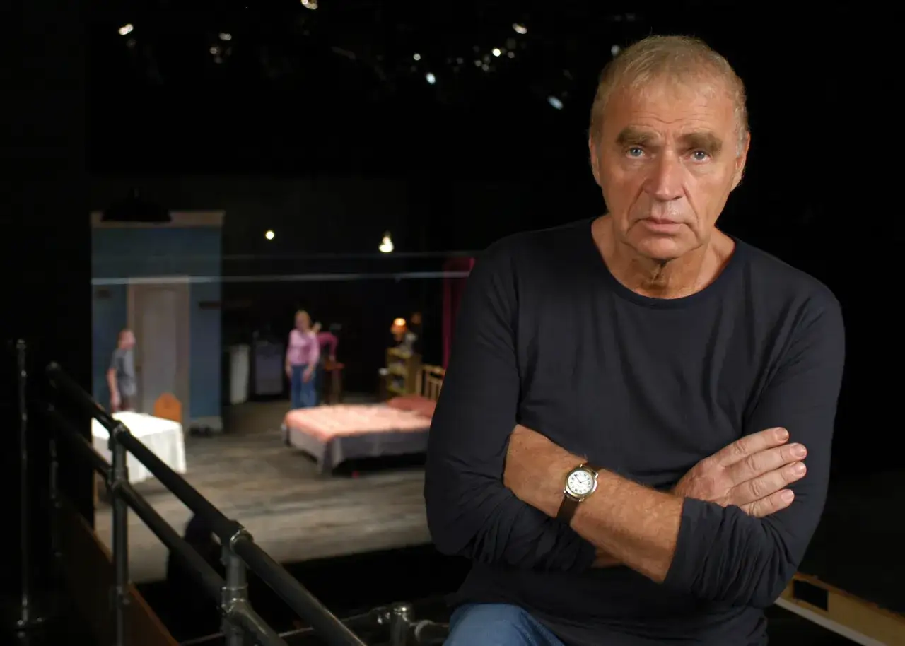 Dawid Głowacki, reżyser teatralny, z rękami skrzyżowanymi na piersi, na tle sceny z łóżkiem i ludźmi.