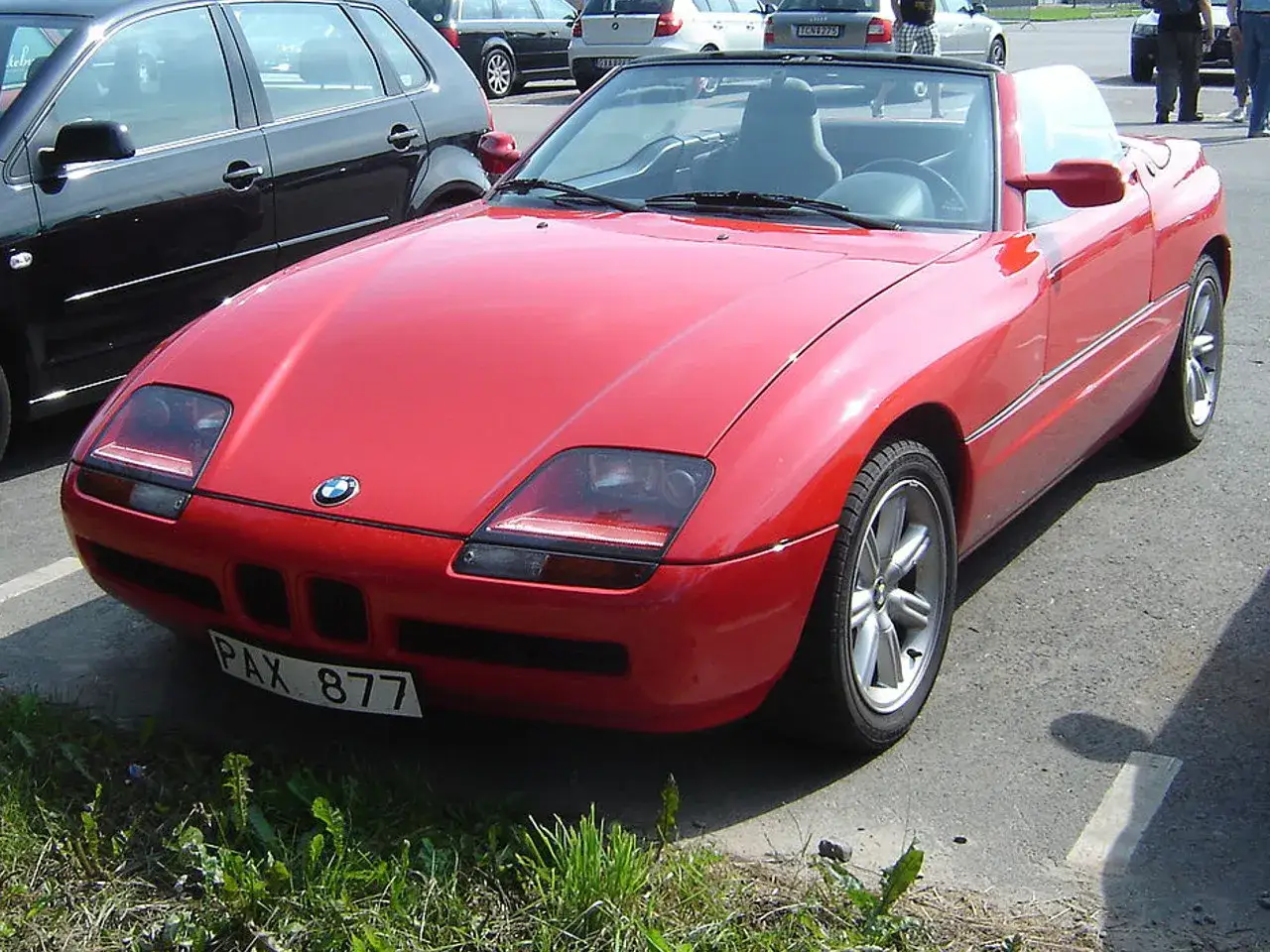 Czerwony kabriolet BMW Z1 z tablicą rejestracyjną PAX 877 zaparkowany obok czarnego samochodu.