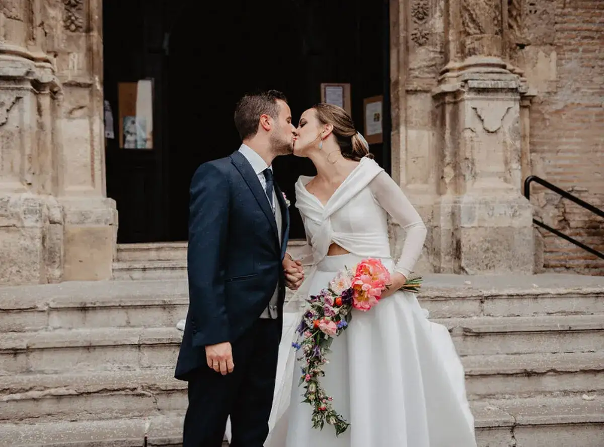 Novios besándose frente a una iglesia, ella lleva un ramo de flores. Un momento especial tras las lecturas de boda religiosa.
