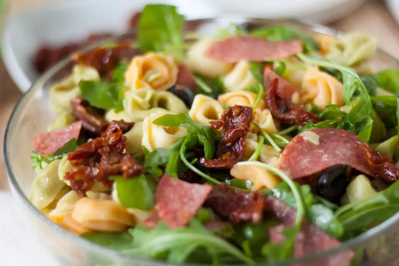 Sałatka z tortellini, suszonymi pomidorami, rukolą i salami w szklanej misce.