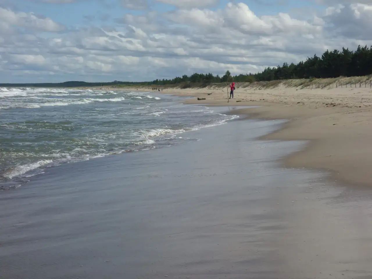 Plaża z falującym morzem i osobą w czerwonej bluzie na piasku.