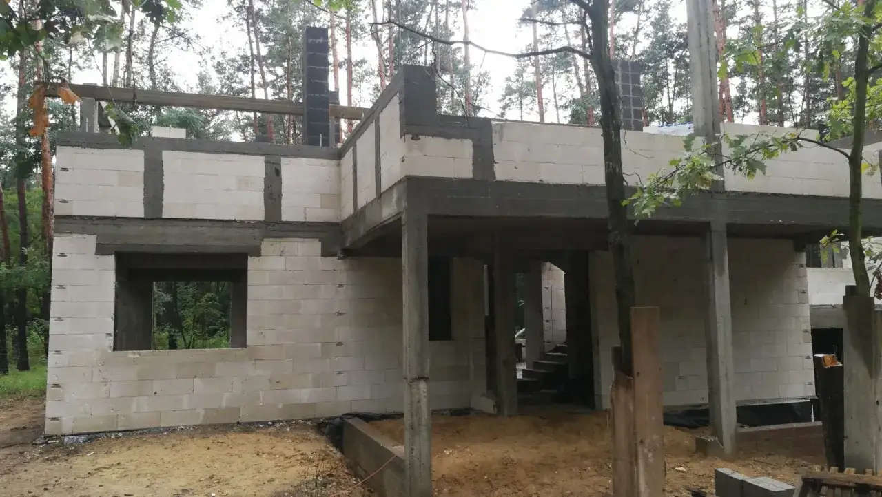 Budowa domu w lesie. Widoczne białe bloczki, betonowe słupy i trzpienie żelbetowe w ścianach murowanych.