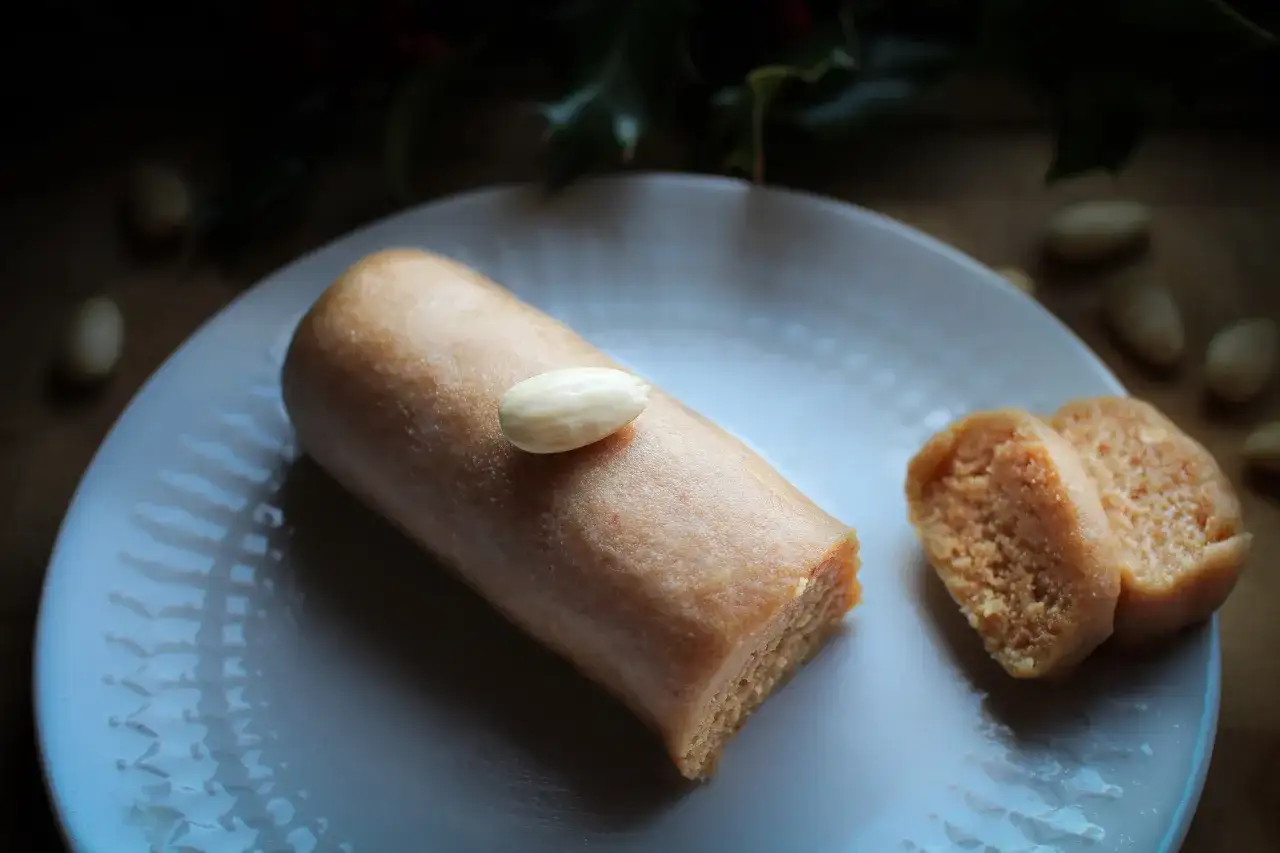 Un bâtonnet de pâte d'amande, garni d'une amande entière, repose sur une assiette blanche. À côté, deux tranches de ce délice, parfait pour un fond de tarte amande.