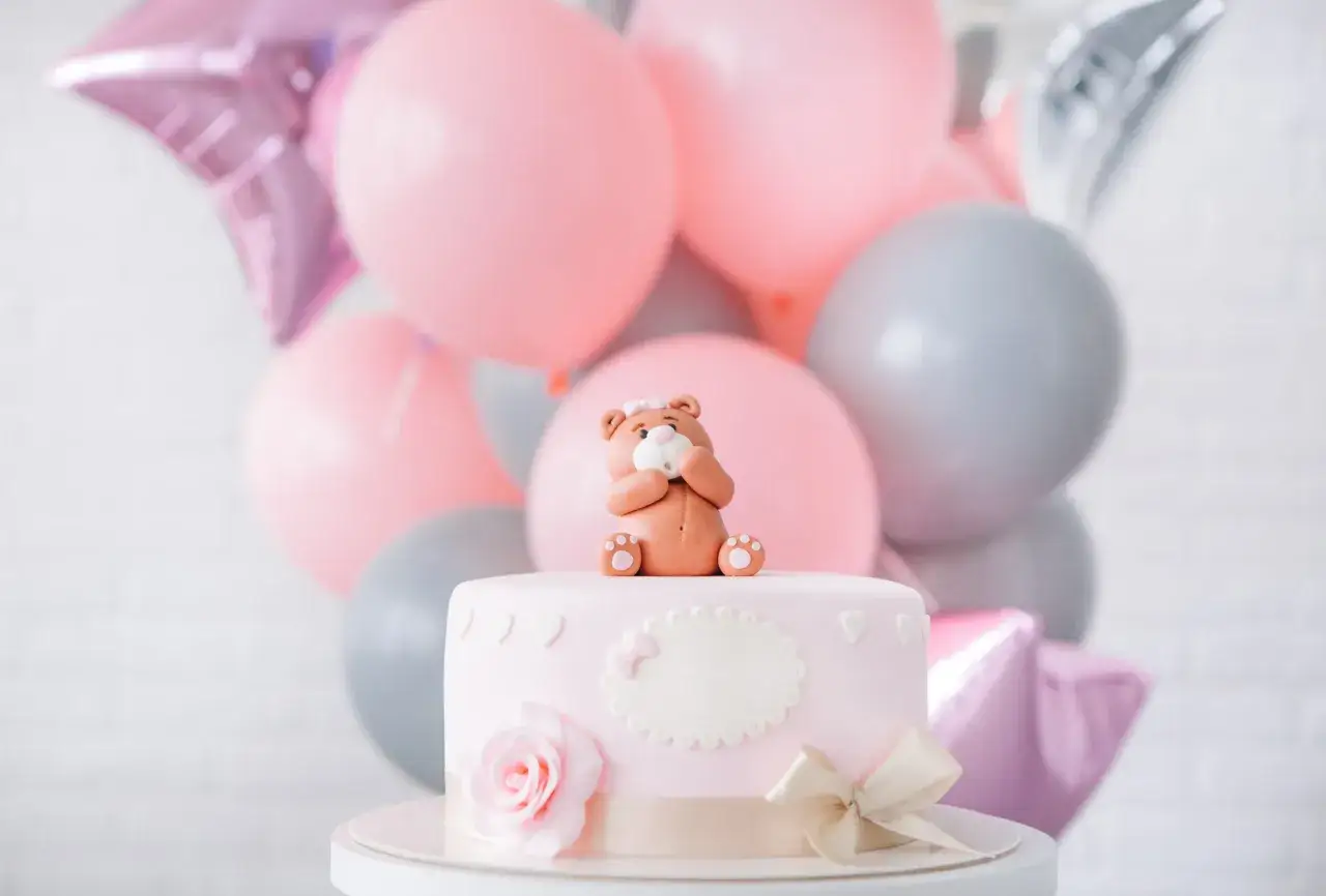 Uroczy topper na tort baby shower w kształcie misia siedzącego na różowym torcie, otoczony balonami w pastelowych kolorach.