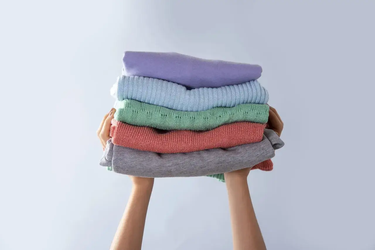 Manos sostienen una pila de ropa doblada en tonos pastel. Consejos para saber como evitar que una prenda destiña.