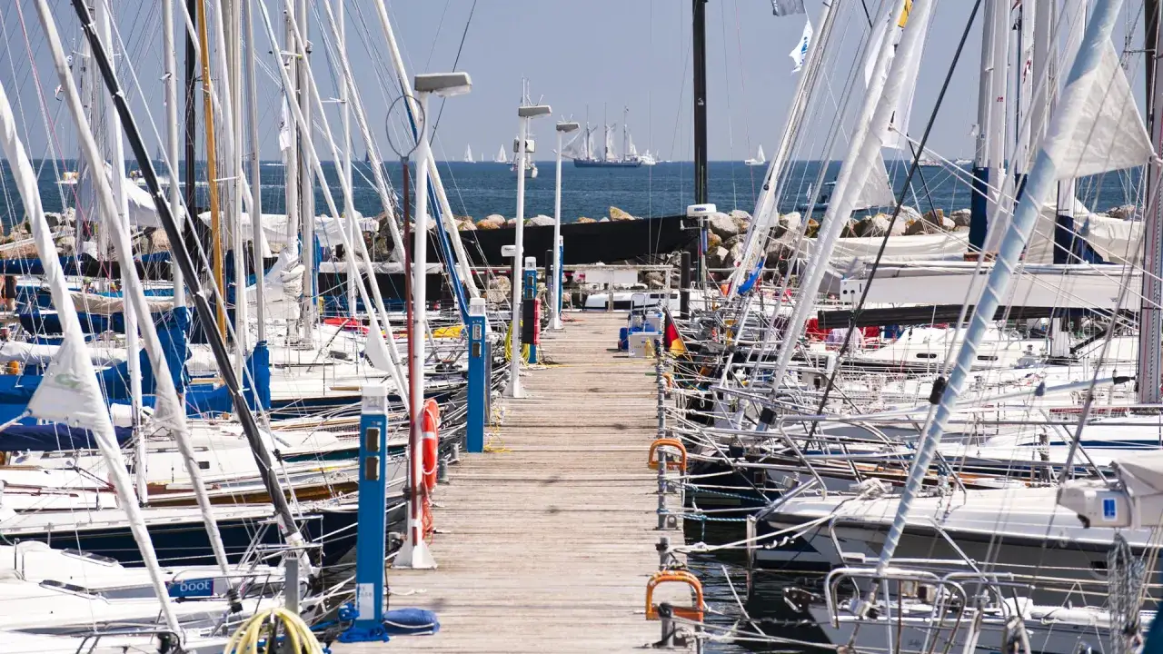 Viele Segelboote liegen im **Flensburg Hafen** an einem Holzsteg. Im Hintergrund segeln große Schiffe auf dem Meer.