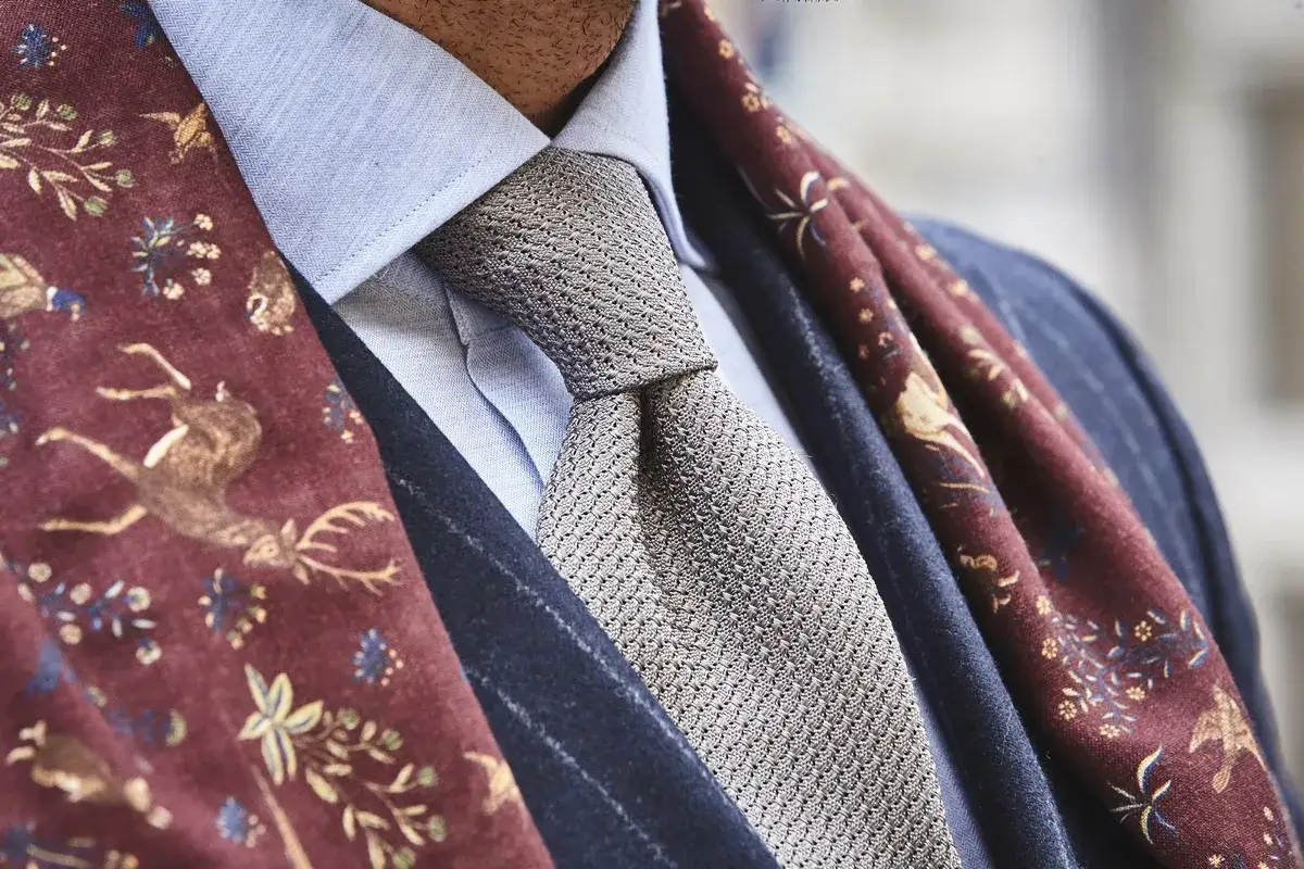 Elegancki mężczyzna w błękitnej koszuli, szarym krawacie i bordowym szalu z motywem jeleni. Całość emanuje nonszalancką elegancją, czyli sprezzaturą.