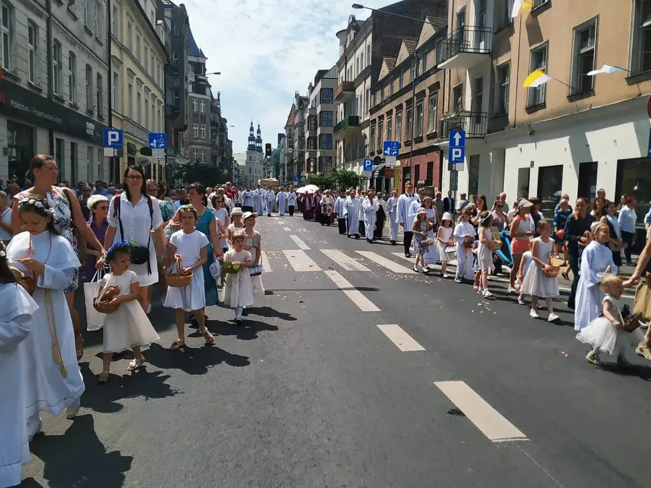 Procesja dzieci w białych strojach, niosących koszyki, idzie ulicą. W tle widać budynki i ludzi, co może symbolizować sekularyzację społeczeństwa.