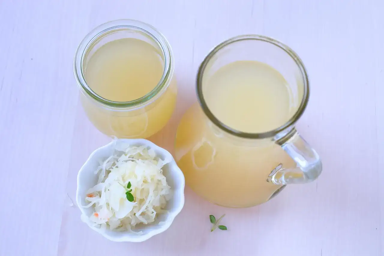 Ein Glas und ein Krug mit Sauerkrautsaft, reich an Vitaminen, stehen neben einer Schale Sauerkraut.