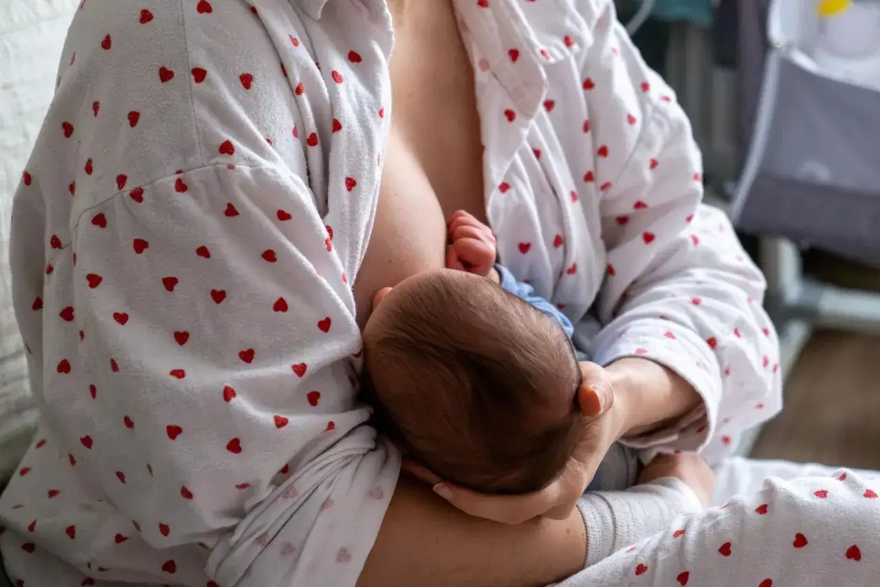 Matka karmi piersią dziecko. W tle widać łóżeczko. Zatkany kanał mleczny to częsty problem.