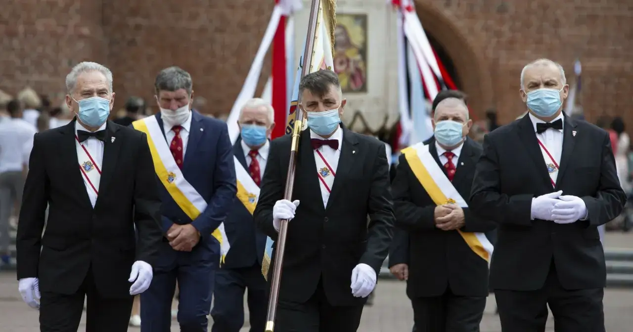 Rycerze Kolumba w garniturach i maskach maszerują z flagą.