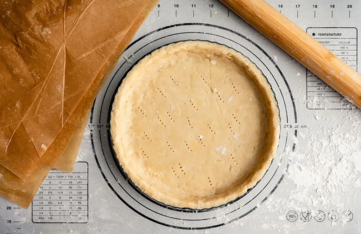 Pâte sucrée noisette prête à être garnie, sur un tapis de pâtisserie avec un rouleau à bois et du papier sulfurisé.