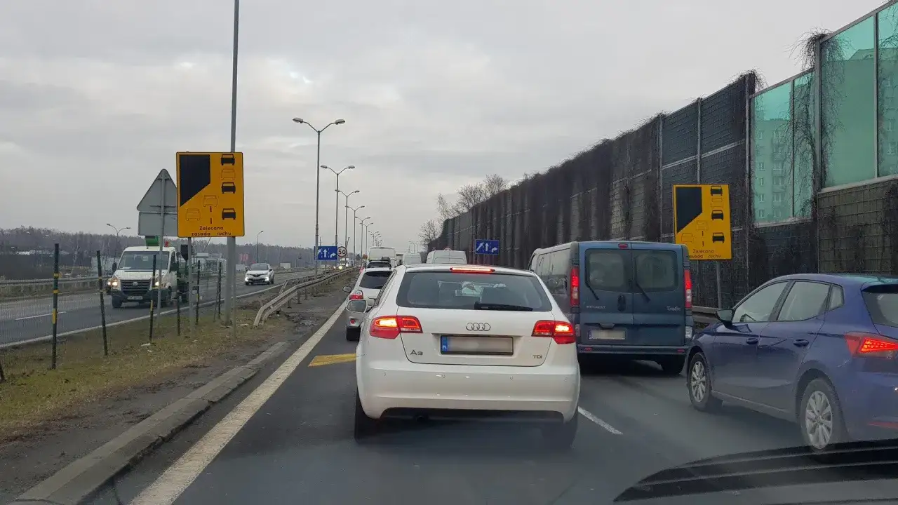 Korek na drodze, samochody stoją w miejscu. Widoczny znak "Zalecana zasada ruchu" informujący o konieczności stosowania jazdy na suwak.