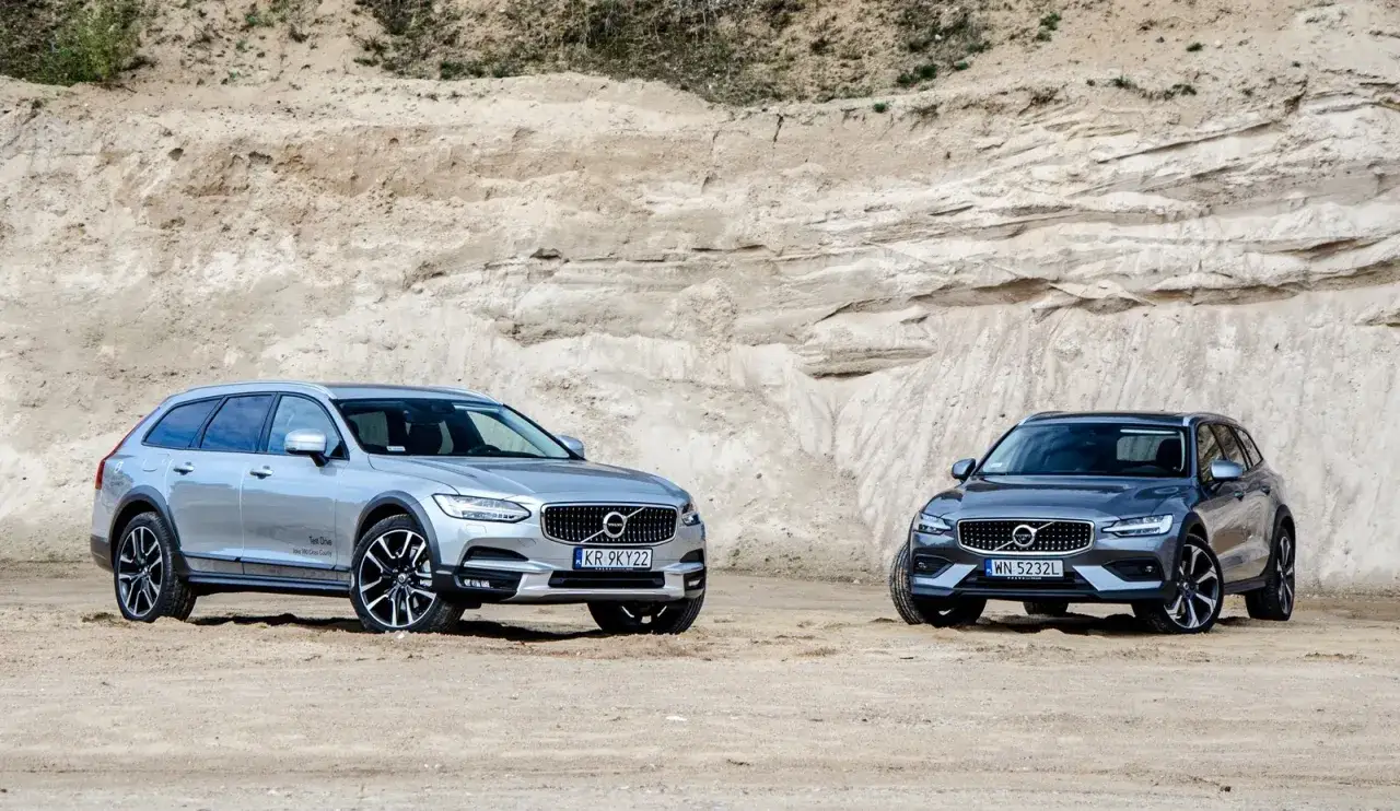Dwa samochody Volvo Cross Country, srebrny i grafitowy, stoją na piaszczystym podłożu przed piaskową ścianą.