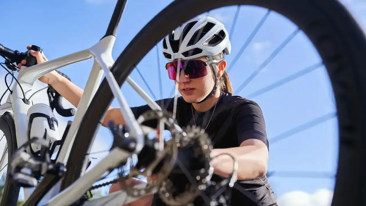 Kobieta w kasku i okularach rowerowych sprawdza rower. Widoczny jest fragment ramy, kierownicy i koła, a także przerzutka.