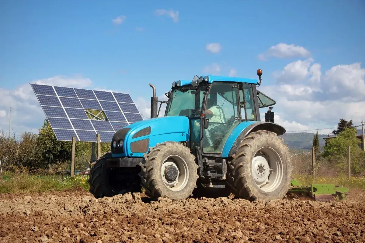 Błękitny traktor pracuje na polu, obok paneli fotowoltaika arimr. Słońce i chmury na niebie.