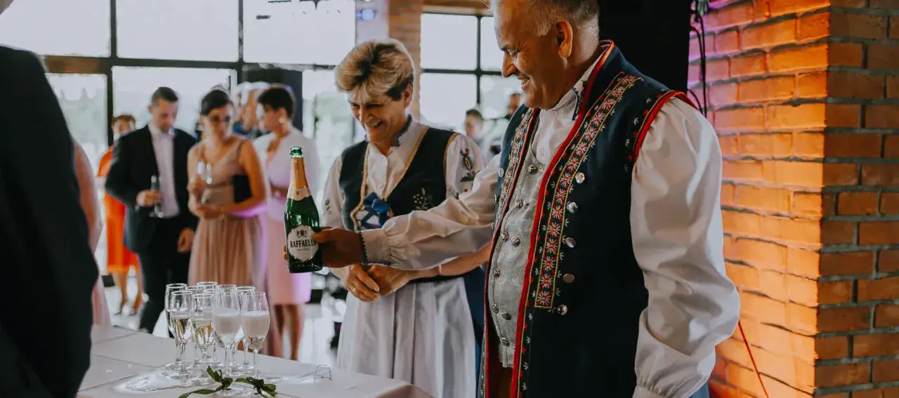 Para w strojach ludowych trzyma butelkę wina musującego na tle weselnych gości.