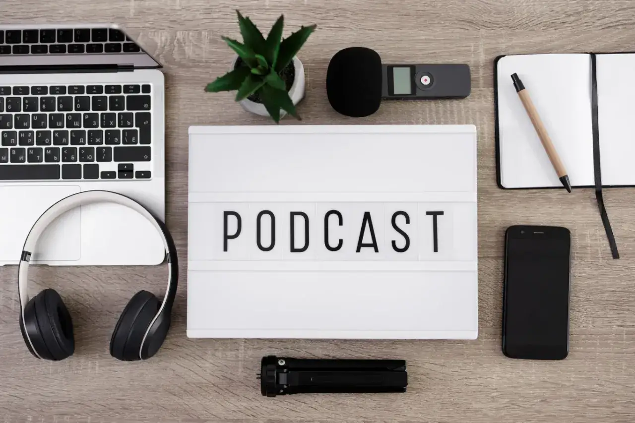 Laptop, słuchawki, mikrofon, dyktafon, notatnik i telefon leżą obok tablicy z napisem PODCAST.