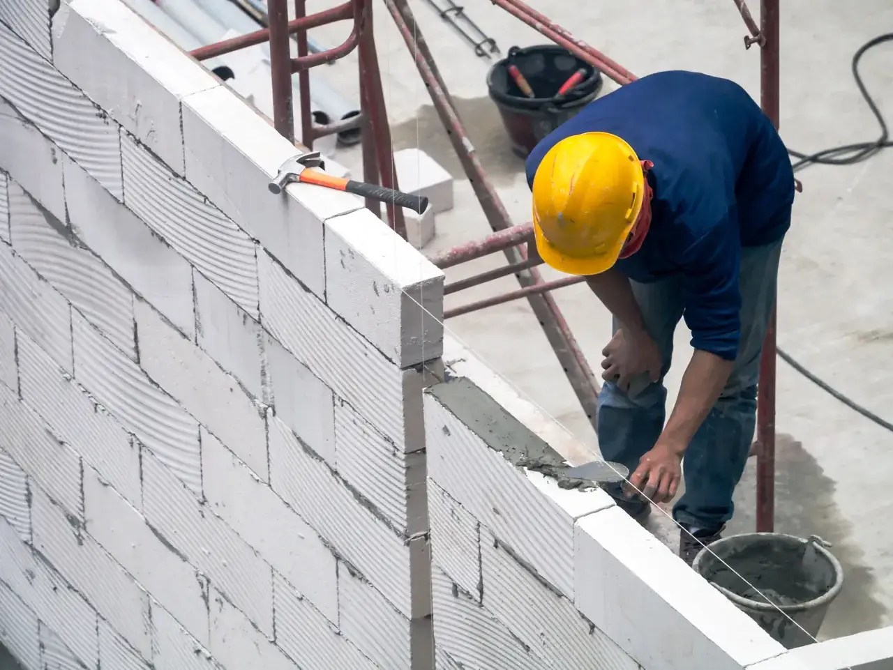 Budowlaniec układa bloczki z betonu komórkowego, dbając o precyzyjne wymiary ściany. Młotek i kielnia czekają na użycie.