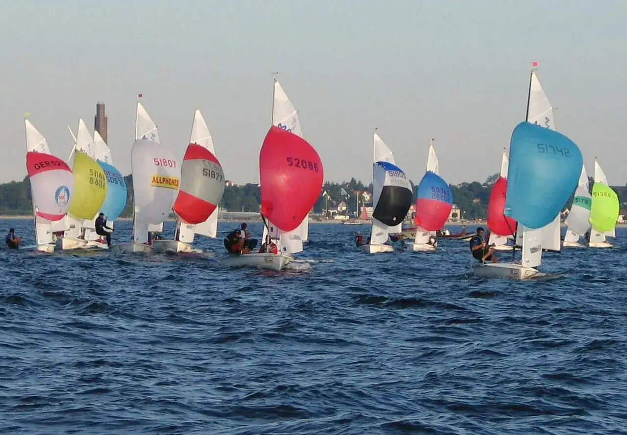 Bunte Segel dominieren das Bild einer spannenden Regatta. Mehrere kleine Boote mit Seglern sind auf dem Wasser unterwegs.