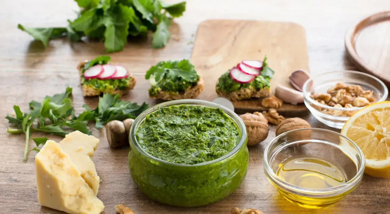 Zielone pesto w misce, obok składniki: ser, orzechy, cytryna, czosnek, oliwa.
