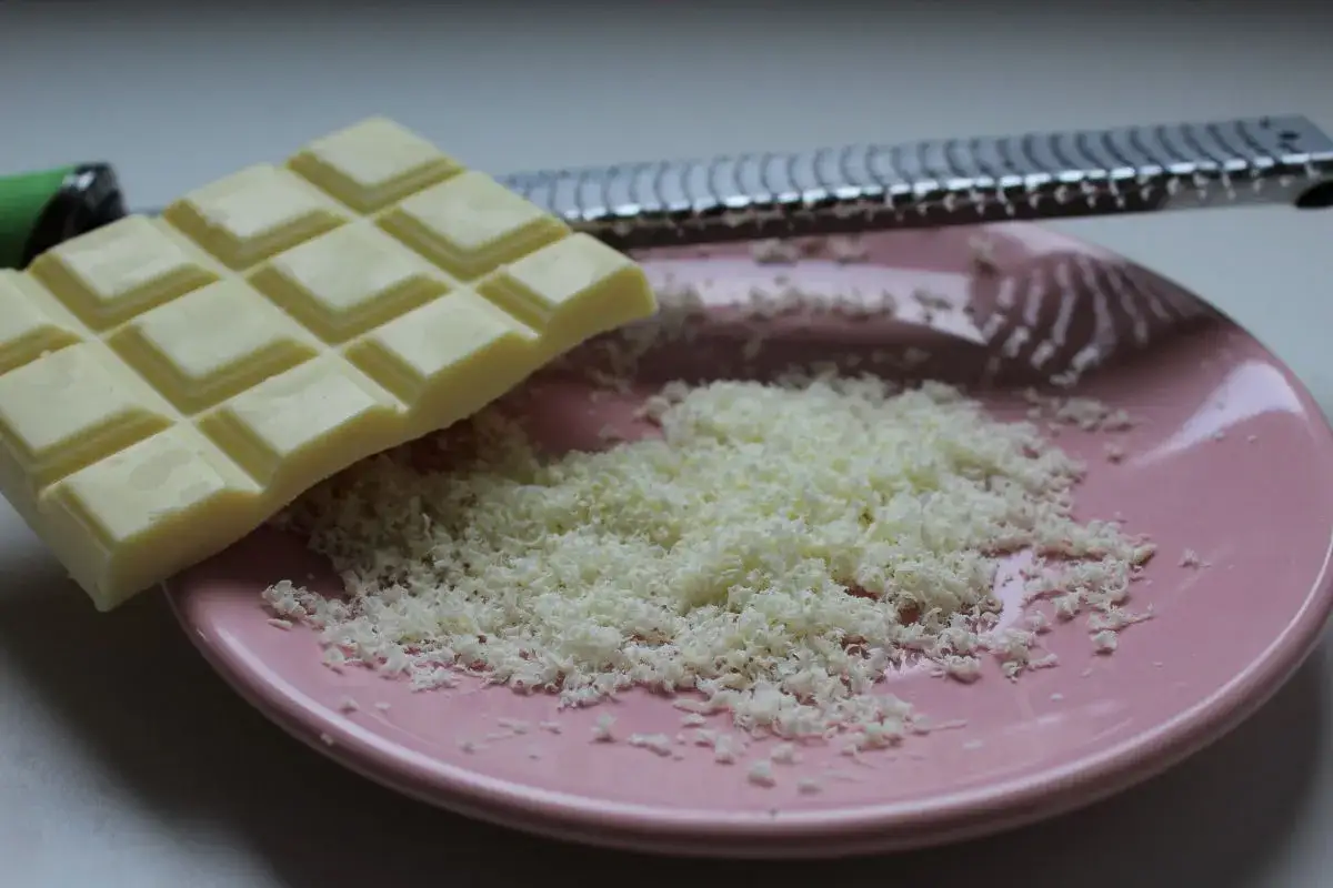 Une râpe à fromage argentée est posée sur une assiette rose, recouverte de copeaux de chocolat blanc fraîchement râpé.