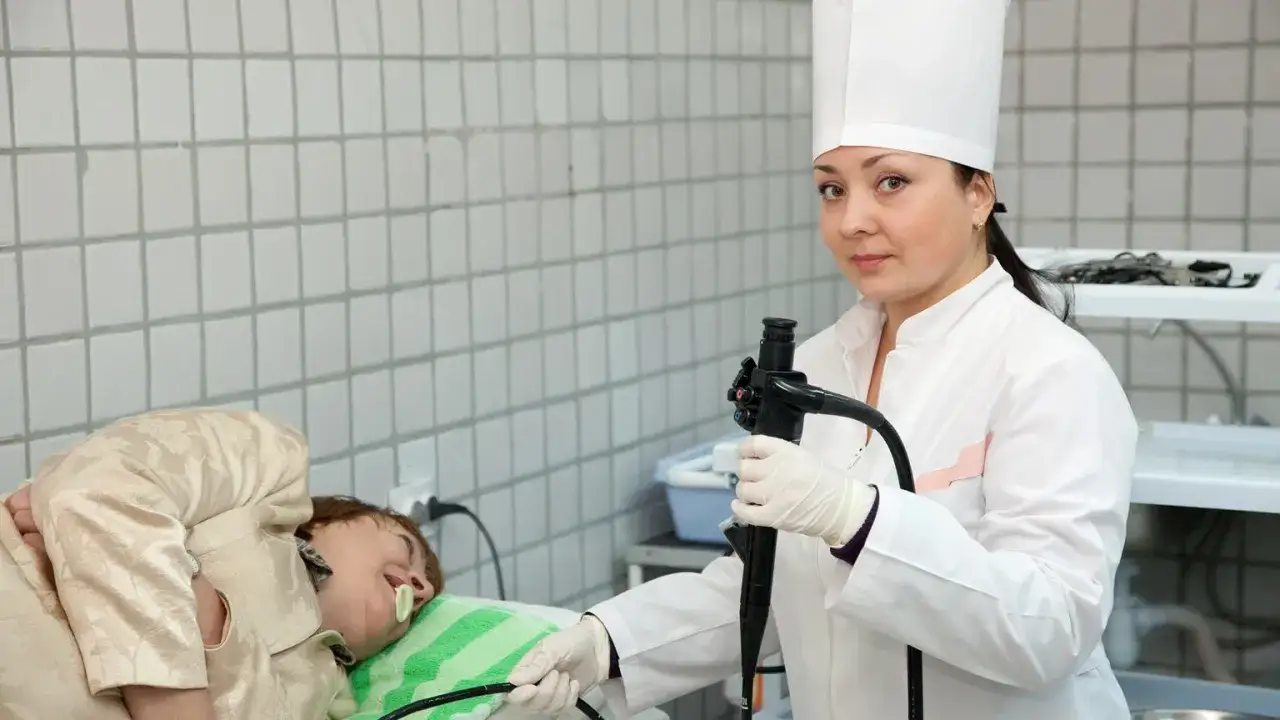 Lekarka w białym fartuchu i czepku przygotowuje pacjenta do gastroskopii, trzymając endoskop.