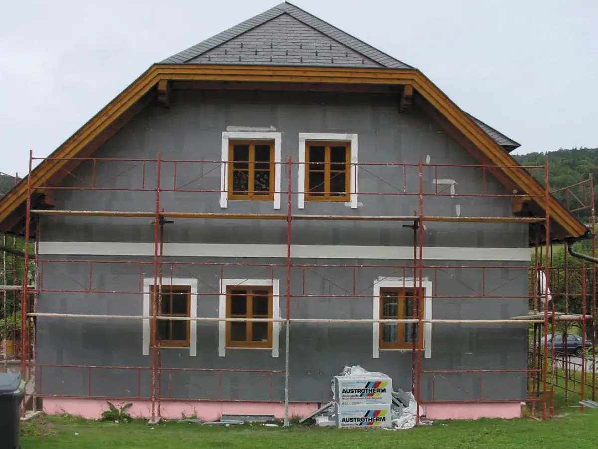 Dom w trakcie ocieplania. Szara fasada, drewniane okna, rusztowanie. Materiały budowlane Austrotherm wskazują na dbałość o niski współczynnik przewodzenia ciepła.
