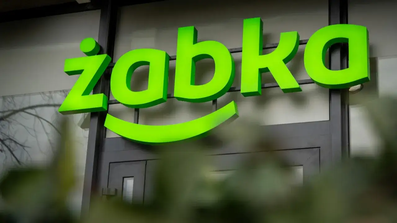 Jasnozielony neonowy napis "Żabka" z uśmiechem pod spodem na tle budynku.