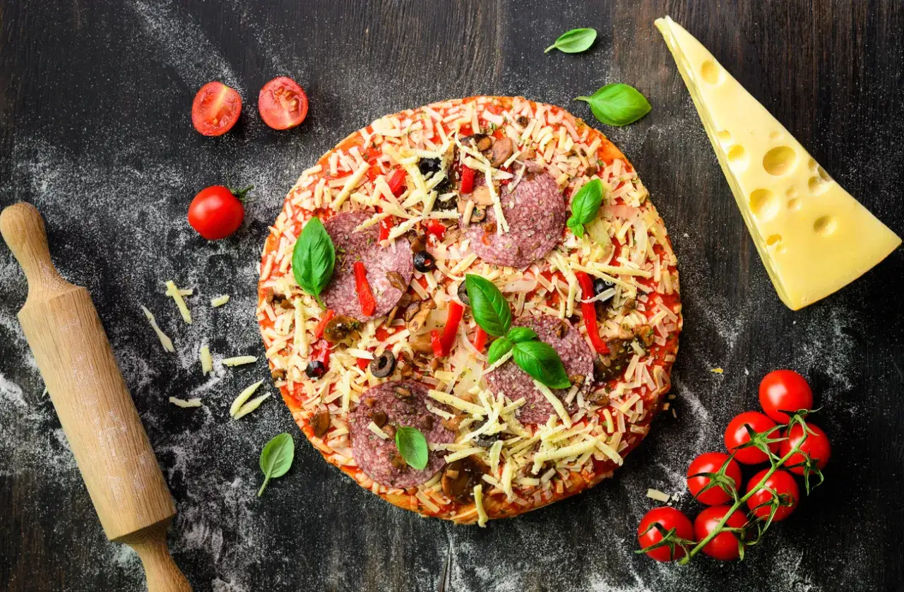Gotowa pizza z salami i warzywami, obok ser, pomidorki i wałek. Idealna na szybki obiad, jaki typ maki na pizze?